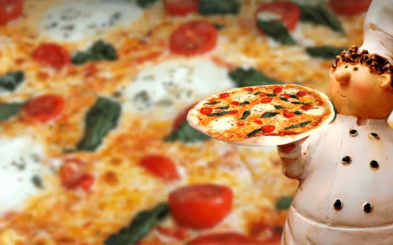 Chef anziano, donna giovane e donna matura cucinano insieme attorno a un pentolino, fra verdure fresche, in cucina luminosa.